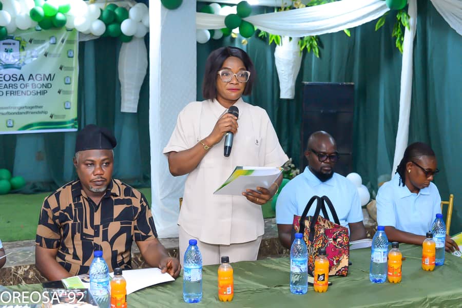 OREOSA92 member standing and addressing an association event with a microphone while holding a document, with three members seated at the table